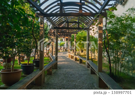 Wooden arched pergola frames a serene garden path lined with stone benches and lush green foliage during a bright day Wooden arched pergola frames a serene garden path lined with stone benches and lush green foliage during a bright day 138997701