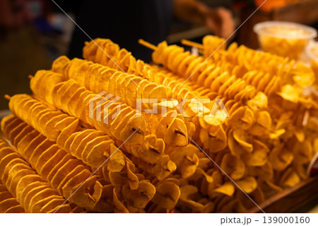 Close-up of crispy, golden spiral-cut tornado potatoes on wooden skewers at a bustling local street food night market. Close-up of crispy, golden spiral-cut tornado potatoes on wooden skewers at a bustling local street food night market. 139000160