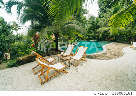 Serene, curved swimming pool surrounded by lush tropical jungle greenery, palm trees, and wooden sun loungers at a sustainable eco-resort. Serene, curved swimming pool surrounded by lush tropical jungle greenery, palm trees, and wooden sun loungers at a sustainable eco-resort. 139000161