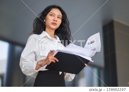 Confident young businesswoman holding a clipboard and financial reports in a modern corporate setting. Professional female manager looking ahead, representing leadership, vision, and success. Confident young businesswoman holding a clipboard and financial reports in a modern corporate setting. Professional female manager looking ahead, representing leadership, vision, and success. 139002297