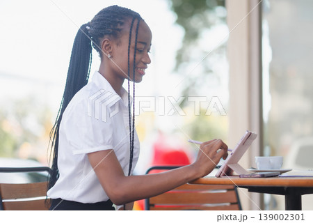 Side view of young Black business woman working on a tablet with stylus at a cafe table. Remote work, digital note taking, online planning, coffee break and modern productivity concept. Side view of young Black business woman working on a tablet with stylus at a cafe table. Remote work, digital note taking, online planning, coffee break and modern productivity concept. 139002301