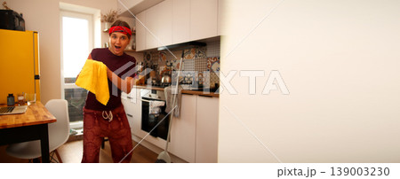 Funny young woman with a bandana holding a yellow microfiber cloth in a modern kitchen. Happy housewife having fun during cleaning. Panoramic view with copy space 139003230