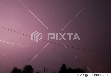 Real lightning strike in the purple night sky during a summer thunderstorm. Dramatic lightning bolt over the silhouettes of trees and power lines. Weather and nature power concept Real lightning strike in the purple night sky during a summer thunderstorm. Dramatic lightning bolt over the silhouettes of trees and power lines. Weather and nature power concept 139003234