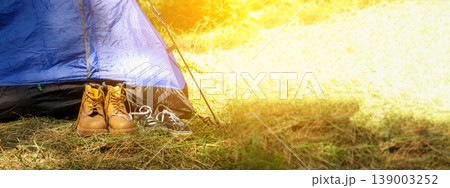 Tourist boots and sneakers standing near a blue tent in a forest. Concept of camping, hiking, and outdoor summer adventures. Panoramic shot with bright sunlight and copy space 139003252