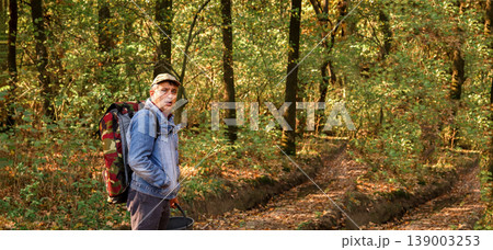 Man with a large backpack and a bucket walking on a dirt road through an autumn forest. Side view of a hiker exploring nature during golden hour. Wide panoramic shot with copy space Man with a large backpack and a bucket walking on a dirt road through an autumn forest. Side view of a hiker exploring nature during golden hour. Wide panoramic shot with copy space 139003253