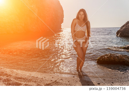 Woman sunset beach standing in shallow ocean waves during golden hour wearing white lingerie on a scenic rocky coastline Woman sunset beach standing in shallow ocean waves during golden hour wearing white lingerie on a scenic rocky coastline 139004736