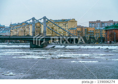 Winter Cityscape With Frozen River and Steel Suspension Bridge Spanning Historic Riverfront Buildings 139006274