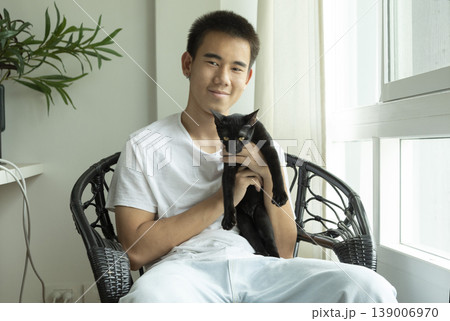 Happy teenage Asian boy holding black cat in white living room. 139006970