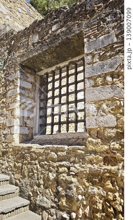 Stone wall displays a barred window with a metal grid, highlighting rustic architecture and historical features in a Mediterranean environment 139007099