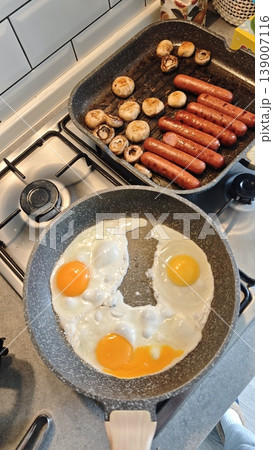 Breakfast preparation with frying pan containing two sunny-side-up eggs, alongside a grill pan with sausages and mushrooms on a modern kitchen stove Breakfast preparation with frying pan containing two sunny-side-up eggs, alongside a grill pan with sausages and mushrooms on a modern kitchen stove 139007116