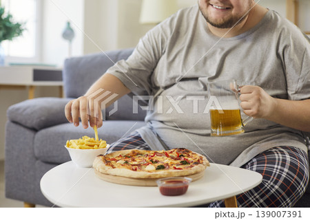 Overweight man holding beer glass mug, eating fries beside pizza at home 139007391