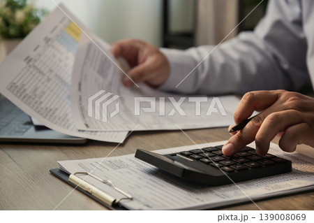 Close up of businessman using calculator and reviewing financial documents on desk, analyzing business report, accounting data, and tax paperwork in modern office, representing finance, audit, and Close up of businessman using calculator and reviewing financial documents on desk, analyzing business report, accounting data, and tax paperwork in modern office, representing finance, audit, and 139008069