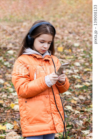 Young girl with headphones using smartphone in autumn park wearing orange jacket and hood Young girl with headphones using smartphone in autumn park wearing orange jacket and hood 139008133