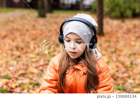 Young girl in orange coat wearing headphones in an autumn park among fallen leaves 139008134