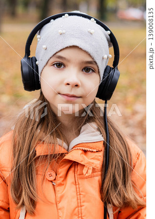 Young girl wearing headphones and an orange jacket listening to music in an autumn park 139008135