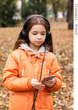 Girl with headphones using smartphone in orange coat outdoors in autumn park 139008152