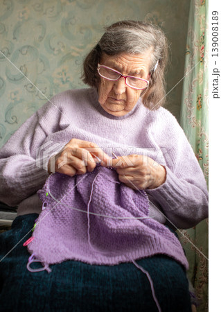 Grandmother knitting a purple sweater at home, focused elderly woman crafting with yarn Grandmother knitting a purple sweater at home, focused elderly woman crafting with yarn 139008189