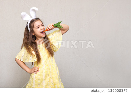 Smiling girl in yellow dress wearing bunny ears and eating a fresh carrot for Easter fun Smiling girl in yellow dress wearing bunny ears and eating a fresh carrot for Easter fun 139008575