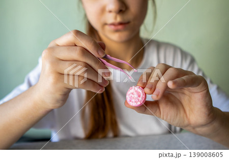 Teen concentrating while using pink tweezers to apply decorative beads to homemade lip balm in jar Teen concentrating while using pink tweezers to apply decorative beads to homemade lip balm in jar 139008605