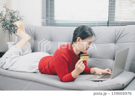 Young Asian woman lying on sofa using laptop and credit card for online shopping at home, digital payment, ecommerce purchase, cashless lifestyle, secure consumer fintech concept indoors 139008976