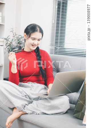 Young Asian woman using laptop and credit card for online shopping at home, browsing ecommerce deals on sofa with shopping bag nearby, digital payment, cashless purchase and retail lifestyle concept Young Asian woman using laptop and credit card for online shopping at home, browsing ecommerce deals on sofa with shopping bag nearby, digital payment, cashless purchase and retail lifestyle concept 139008977