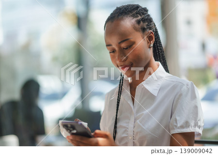 Young African woman using smartphone while holding coffee cup in a cafe, concept of mobile lifestyle, online communication, remote work, digital routine and modern casual business. 139009042