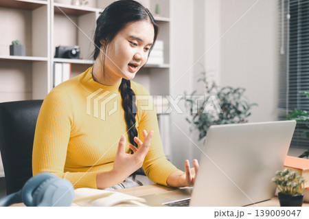 Young Asian woman speaking during online class on laptop at home office desk, learning remotely with expressive hand gestures. Webinar, video call, e-learning, communication and study concept. Young Asian woman speaking during online class on laptop at home office desk, learning remotely with expressive hand gestures. Webinar, video call, e-learning, communication and study concept. 139009047