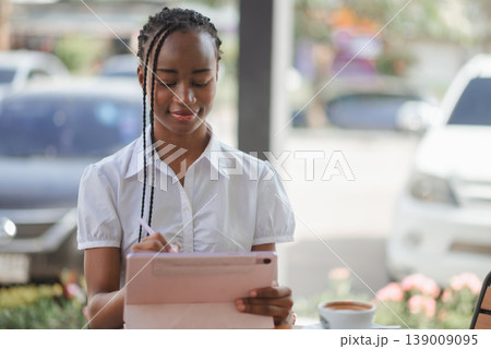 Young African business woman writing on tablet with stylus at outdoor cafe table with coffee, concept of digital note taking, planning, remote work and modern paperless productivity. 139009095
