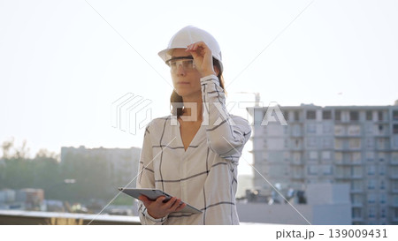 Woman construction engineer wearing white checked blouse and hard hat is using tablet computer while inspecting a building site at sunset, front view. Architecture and engineering concepts 139009431