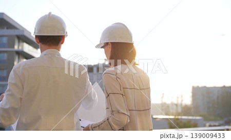 Man and woman architects, wearing safety helmets and glasses, are examining blueprints at a construction site during a sunset, back view. Architecture concept Man and woman architects, wearing safety helmets and glasses, are examining blueprints at a construction site during a sunset, back view. Architecture concept 139009433