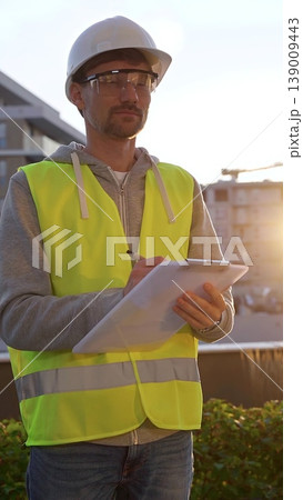 Man engineer with white helmet and safety vest is taking notes on a clipboard while inspecting a construction site at sunrise in early morning, vertical front view. Architecture and engineering Man engineer with white helmet and safety vest is taking notes on a clipboard while inspecting a construction site at sunrise in early morning, vertical front view. Architecture and engineering 139009443