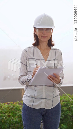 Woman a construction engineer wearing white checked blouse and hard hat is making notes on a clipboard while inspecting a building site in foggy weather, front vertical view. Architecture concept Woman a construction engineer wearing white checked blouse and hard hat is making notes on a clipboard while inspecting a building site in foggy weather, front vertical view. Architecture concept 139009444