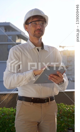Male construction engineer wearing white shirt and helmet is using digital tablet while inspecting a building site at sunrise, front and vertical view . Architecture concept Male construction engineer wearing white shirt and helmet is using digital tablet while inspecting a building site at sunrise, front and vertical view . Architecture concept 139009446