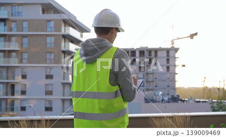 Man engineer with a white protective helmet and safety vest is writing on a clipboard while inspecting a construction site at sunset. Back view Man engineer with a white protective helmet and safety vest is writing on a clipboard while inspecting a construction site at sunset. Back view 139009469