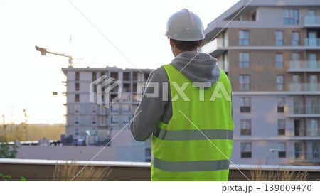 Man engineer with a white protective helmet and safety vest is writing on a clipboard while inspecting a construction site in early morning at sunrise. Architect concept Man engineer with a white protective helmet and safety vest is writing on a clipboard while inspecting a construction site in early morning at sunrise. Architect concept 139009470