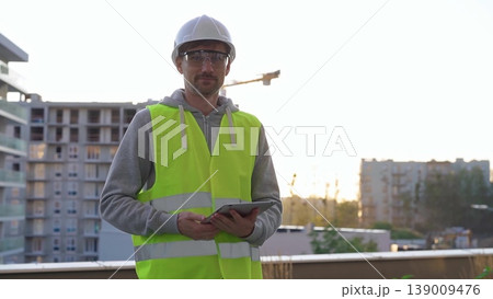 Man construction site engineer holding tablet and wearing hard hat and safety vest with a sunrise at the background in early morning Man construction site engineer holding tablet and wearing hard hat and safety vest with a sunrise at the background in early morning 139009476