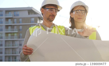Architects or engineers wearing safety helmets and vests holding blueprint and discussing something on construction site at sunrise, front view. Architecture and engineering Architects or engineers wearing safety helmets and vests holding blueprint and discussing something on construction site at sunrise, front view. Architecture and engineering 139009477