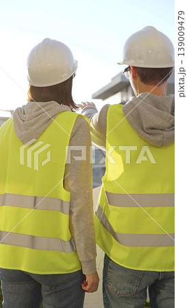 Man and woman architects or engineers wearing safety hard hats and vests holding blueprint and discussing something on construction site at sunrise, back vertical view. Architecture concept Man and woman architects or engineers wearing safety hard hats and vests holding blueprint and discussing something on construction site at sunrise, back vertical view. Architecture concept 139009479