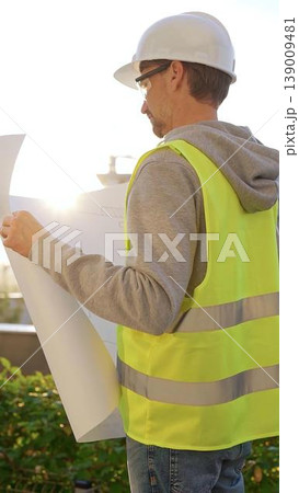 Male architect wearing safety hard hat and vest holding blueprint and looking to construction site at sunrise, back vertical view. Architecture and engineering Male architect wearing safety hard hat and vest holding blueprint and looking to construction site at sunrise, back vertical view. Architecture and engineering 139009481