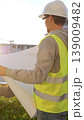 Man an architect or engineer wearing safety hard hat and vest holding blueprint and looking to construction site at sunrise, back vertical view. Architecture and engineering 139009482