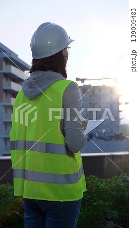 Woman construction engineer with a white hard hat and safety vest is using digital tablet while inspecting a construction site in the early morning at sunrise, back view Woman construction engineer with a white hard hat and safety vest is using digital tablet while inspecting a construction site in the early morning at sunrise, back view 139009483