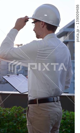 Man a constructive engineer wearing white shirt and hard hat is making notes on a clipboard while inspecting a building site at sunrise, back and vertical view . Architect at work Man a constructive engineer wearing white shirt and hard hat is making notes on a clipboard while inspecting a building site at sunrise, back and vertical view . Architect at work 139009486
