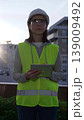 Female architect or engineer wearing safety hard hat and vest holding digital tablet while inspecting a construction site at sunrise in early morning, front vertical view. Architecture concept 139009492