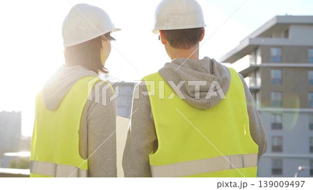 Two architects wearing safety hard hats and vests holding blueprint and discussing construction site project at sunrise, back view. Architecture, engineering and teamwork concept Two architects wearing safety hard hats and vests holding blueprint and discussing construction site project at sunrise, back view. Architecture, engineering and teamwork concept 139009497
