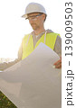 Man an architect or engineer wearing safety hard hat and vest holding blueprint and looking to construction site at sunrise, front vertical view. Architecture concept 139009503