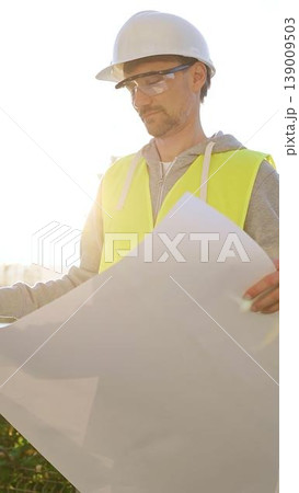 Man an architect or engineer wearing safety hard hat and vest holding blueprint and looking to construction site at sunrise, front vertical view. Architecture concept Man an architect or engineer wearing safety hard hat and vest holding blueprint and looking to construction site at sunrise, front vertical view. Architecture concept 139009503