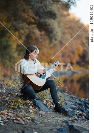 Portrait of a young girl playing the guitar near river. Bright autumn trees on the background, practicing playing the guitar Portrait of a young girl playing the guitar near river. Bright autumn trees on the background, practicing playing the guitar 139011403
