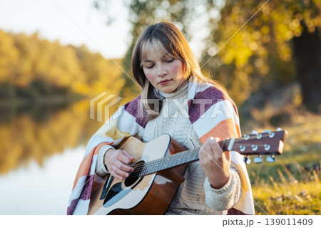 Young girl playing the guitar near the river. Female playing the guitar in bright autumn park sitting on the river bank 139011409