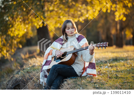 Portrait of a young girl playing the guitar outdoors. Beautiful girl enjoying fall nature, playing the guitar alone in autumn park Portrait of a young girl playing the guitar outdoors. Beautiful girl enjoying fall nature, playing the guitar alone in autumn park 139011410