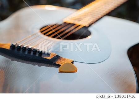Guitar with a plectrum against setting evening sun. Close up of yellow plectrum on a new guitar outdoors Guitar with a plectrum against setting evening sun. Close up of yellow plectrum on a new guitar outdoors 139011416